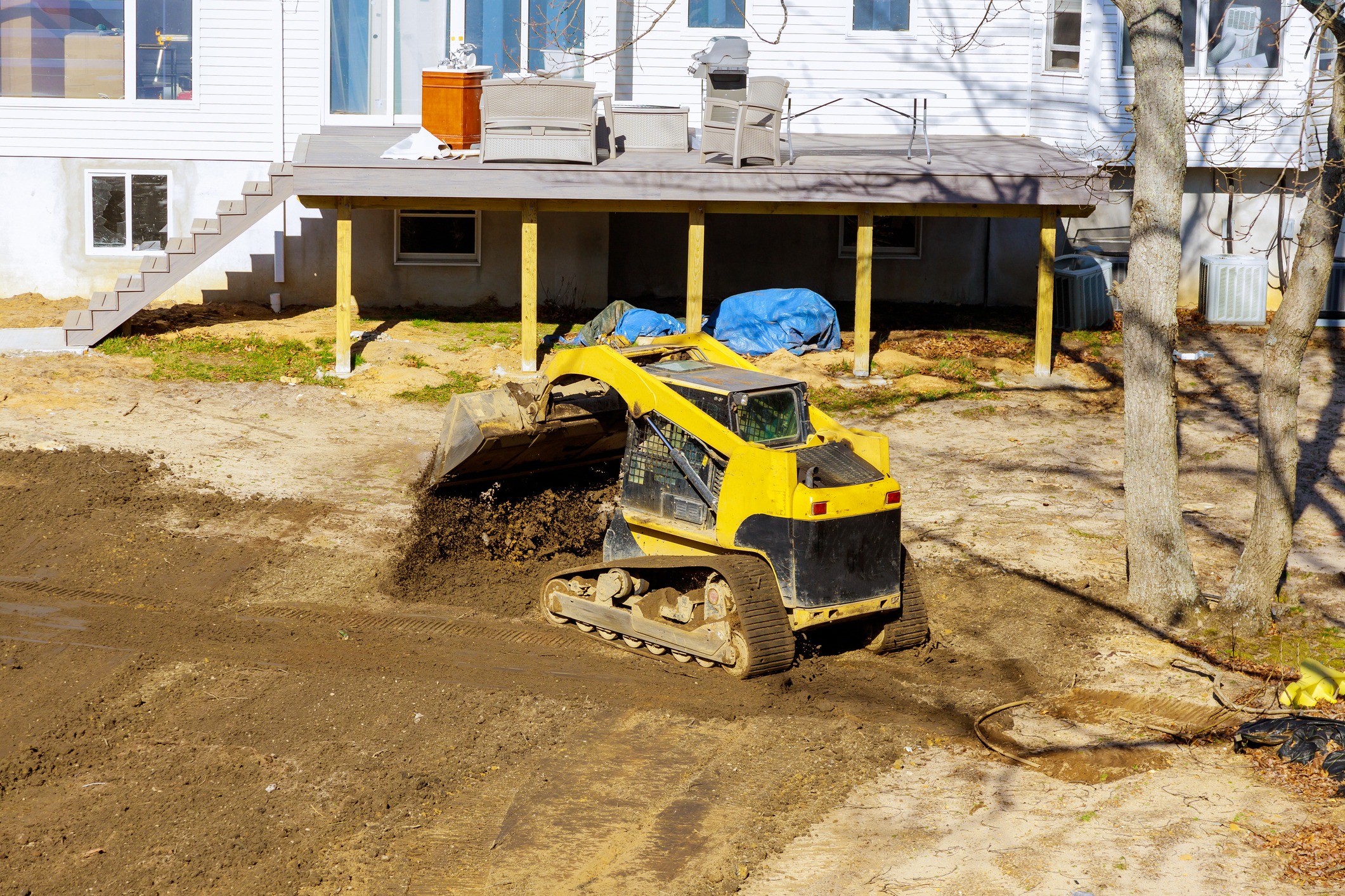 Yellow skid-steer grading backyard beneath raised deck